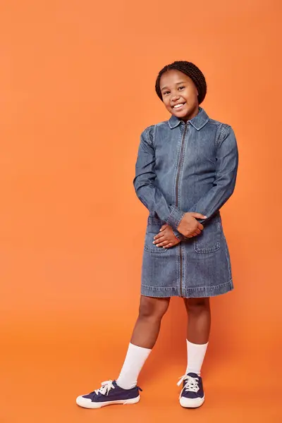 オレンジ色の背景に立っているデニムのドレスで陽気なアフリカ系アメリカ人の少女の全長 — ストック写真