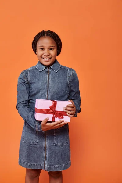 stock image cheerful african american girl in denim dress holding present and looking at camera on orange