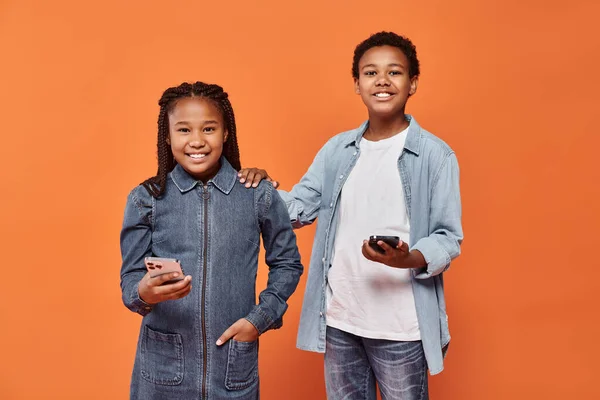 stock image happy african american girl and boy in casual wear holding smartphones on orange background