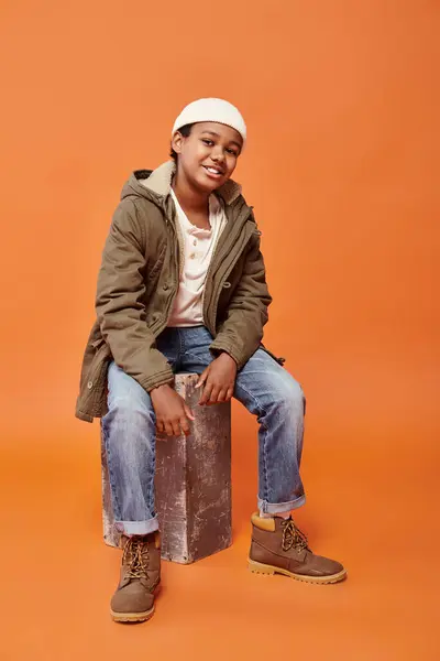 jolly african american boy in winter jacket and jeans with beanie hat sitting and smiling at camera