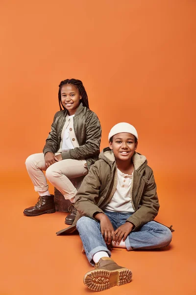 Stock image cheerful preteen african american boy and girl in winter attire sitting on floor on ornage backdrop