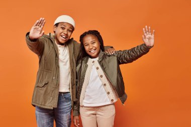 happy african american girl and boy in winter outfits joyfully posing together on orange backdrop clipart