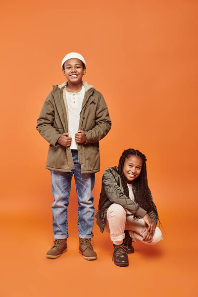 веселые юные африканские американские друзья в теплых куртках позируют в движении на оранжевом фоне