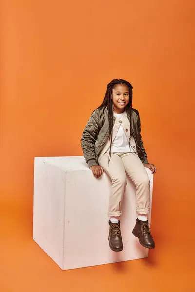 stock image happy african american girl in winter attire smiling at camera on white cube on orange backdrop