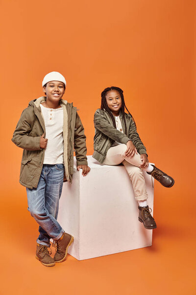happy preteen african american friends in winter outfits smiling ta camera next to white cube