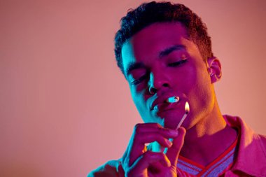 Afro-Amerikalı bir adam pembe arka planda mavi neon ışıkların altında kibritle sigara yakıyor.