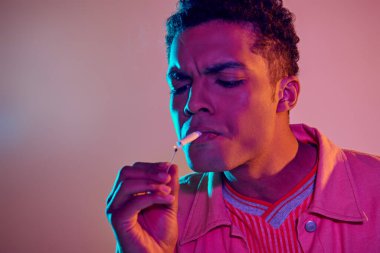 Odaklanmış Afro-Amerikalı adam pembe arka planda mavi neon ışıkların altında kibritle sigara yakıyor.