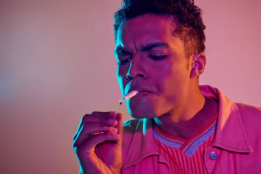 focused african american man lighting cigarette with match under blue neon lights on pink background clipart