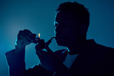 curly african american man holding lighter and smoking pipe on dark blue backdrop with lighting clipart