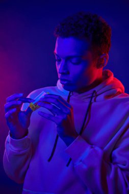 curly african american man rolling cigarette while standing on dark blue background with neon light clipart