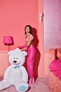 Pembe bluzlu ve pantolonlu neşeli genç bir kadın canlı odada dev ayıcığın yanında gülüyor.