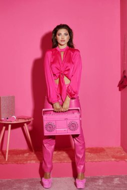 Canlı giyinmiş genç bir kadın elinde retro teyp ile pembe arka planda kameraya bakıyor.