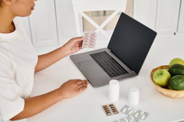 cropped african american nutritionist with pills looking at laptop during an online consultation clipart