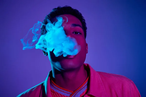 stock image young african american man exhaling smoke against blue background with purple lighting, gen z