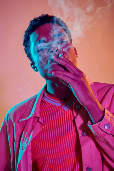 stock image cool and young african american man smoking cigarette under blue neon lights on pink background