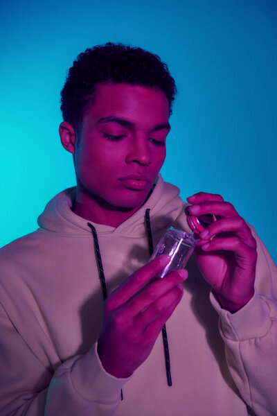 curly african american man in hoodie holding jar with medical cannabis on blue background