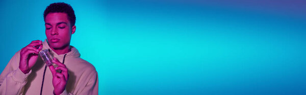 young african american man in hoodie holding jar with medical cannabis on blue background, banner