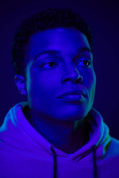 handsome young african american man in hoodie looking away under blue neon light, male beauty