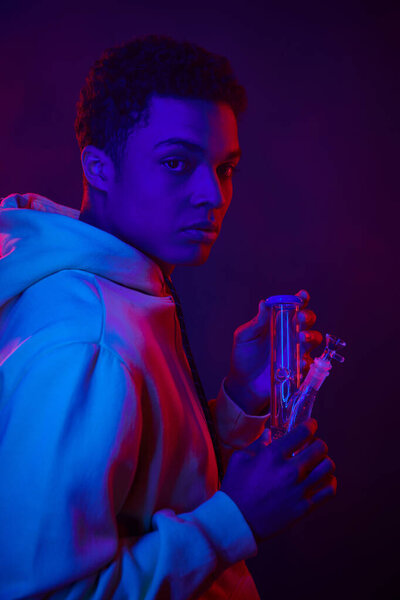 молодой африканский американец в толстовке смотрит на стеклянный бонг на темно-синем фоне с освещением