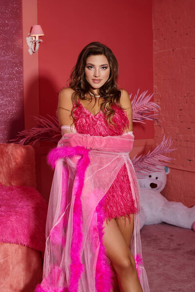 гламурная молодая женщина в розовом пернатом халате и перчатках позирует рядом с плюшевым мишкой на размытом фоне