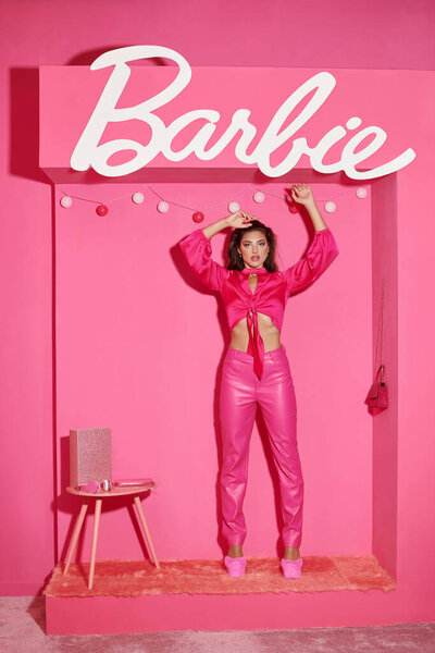 KYIV, UKRAINE - JULY 14, 2023:glamorous young woman in crop top and pink pants standing with raised hands near Barbie letters