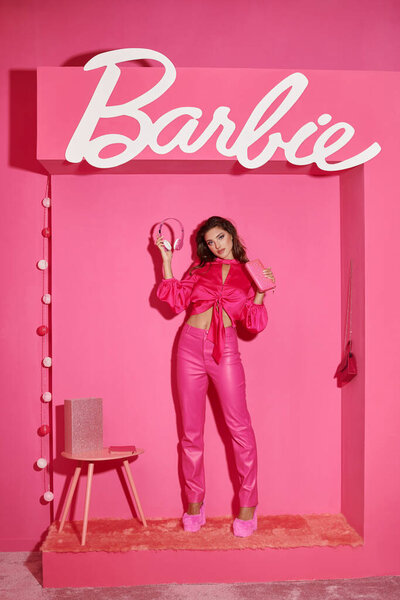 KYIV, UKRAINE - JULY 14, 2023:beautiful doll like woman in crop top and pink pants standing with headphones near Barbie sign
