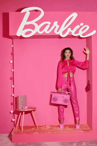 stock image KYIV, UKRAINE - JULY 14, 2023:beautiful young woman in pink attire holding retro boombox and waving hand near Barbie sign