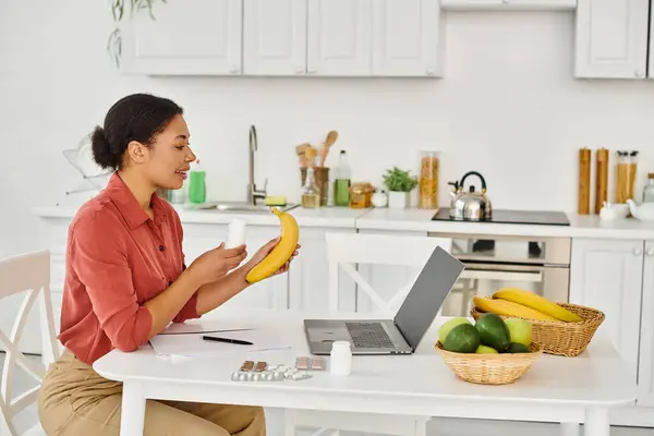 stock image happy african american nutritionist holding banana and giving diet advice on laptop in kitchen