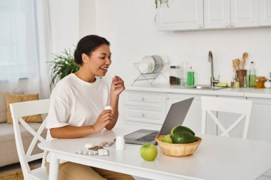 happy african american nutritionist taking medication in front of laptop near pills and fruits clipart