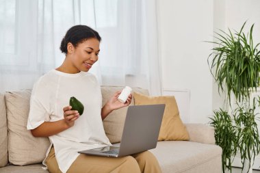 happy african american nutritionist with supplements and avocado giving dietary advice via laptop clipart