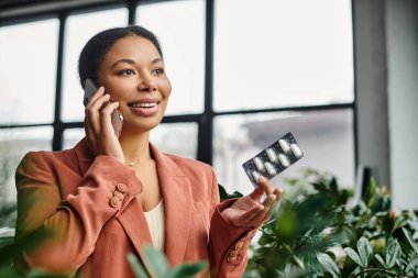joyful african american nutritionist consulting via smartphone while holding supplements in office clipart