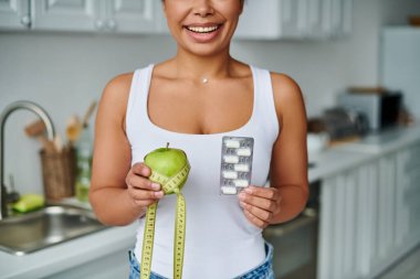 Kırpılmış mutlu Afro-Amerikalı kadın elma ve takviyeli şeritle ölçüm yapıyor.