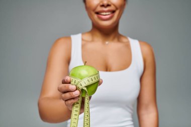 cropped view of happy african american woman with measuring tape and apple on grey background clipart