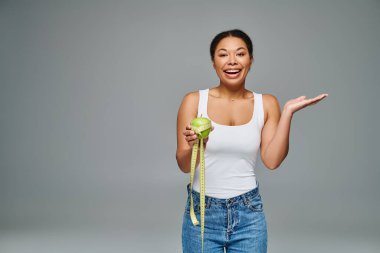 portrait of happy african american woman with measuring tape and apple on grey background clipart