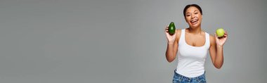 Elma ve avokadoyla ışıl ışıl Afro-Amerikalı kadın gri pankartta dengeli beslenmeyi teşvik ediyor.