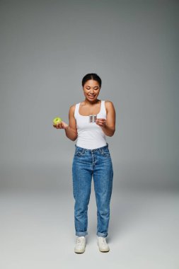 Mutlu Afro-Amerikan kadını yeşil elma grisi arka planla vitaminleri karşılaştırıyor.
