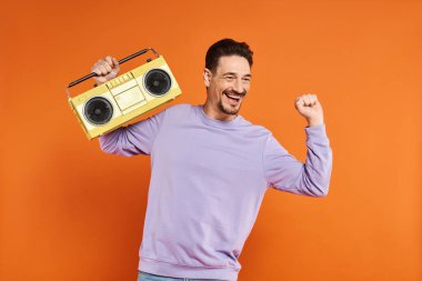 Mor kazaklı neşeli ve sakallı adam elinde turuncu arka planda retro boombox, müzik