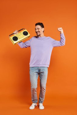 Heyecanlı ve sakallı mor kazaklı adam elinde turuncu arka planda retro boombox, müzik