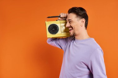Mor kazaklı neşeli adam elinde retro teyp ve turuncu arka planda müzik dinliyor.