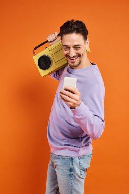 neşeli ve sakallı bir adam akıllı telefon kullanıyor ve elinde turuncu arka planda retro boombox tutuyor, müzik