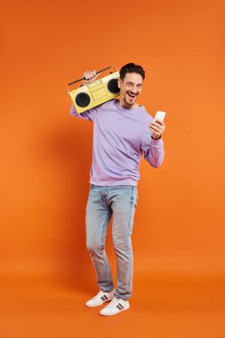 neşeli ve sakallı bir adam akıllı telefon kullanıyor ve elinde turuncu arka planda retro boombox tutuyor, müzik