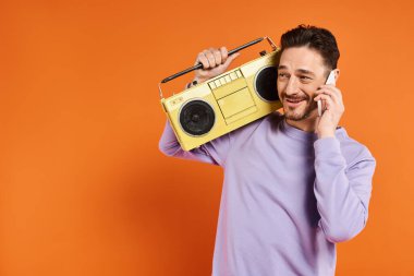 Neşeli ve sakallı bir adam akıllı telefondan konuşuyor ve elinde turuncu arka planda retro teyp var.