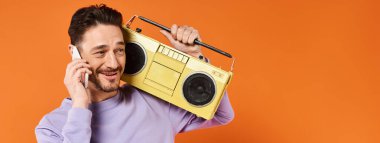 Neşeli ve sakallı bir adam akıllı telefondan konuşuyor ve elinde turuncu arka planda retro teyp var.