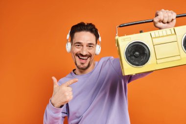 Mor kazaklı ve kablosuz kulaklıklı sakallı mutlu adam turuncu arka planda teyp tutuyor.