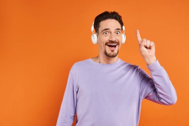 Kablosuz kulaklıklı sakallı adam turuncu arka planda müzik dinliyor, fikir jesti yapıyor.