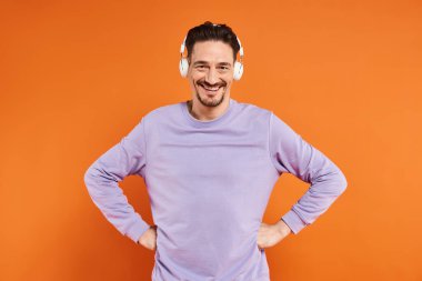 Mor kazaklı ve kablosuz kulaklıklı neşeli adam turuncu arka planda müzik dinliyor.