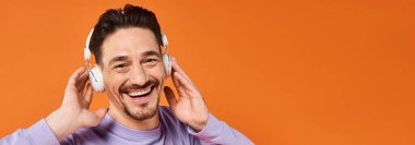 Mor kazaklı, kablosuz kulaklıklı turuncu arka planda müzik dinleyen pozitif bir adam.