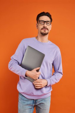Gözlüklü, mor kazaklı, dizüstü bilgisayarlı ve turuncu arka planda duran sakallı bir adam.