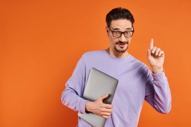 Gözlüklü sakallı adam ve elinde dizüstü bilgisayarla turuncu arka planı işaret ediyor.