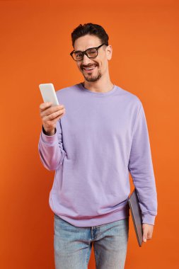 Gözlüklü ve mor kazaklı mutlu adam turuncu arka planda akıllı telefon kullanıyor, mesajlaşıyor.